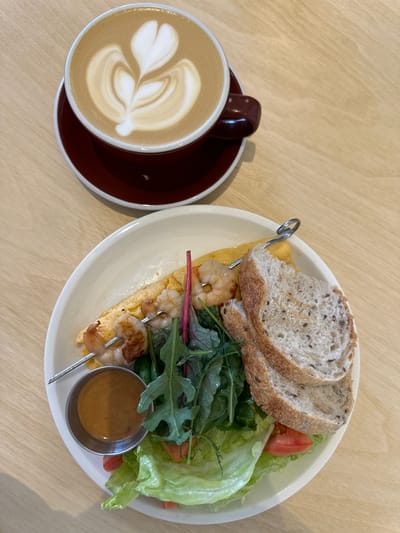 A top-down, close-up shot captures a meal and a coffee cup on a light-colored wooden table. To the left, a dark red coffee cup with a white rim holds a latte with elaborate foam art. The cup rests on a matching saucer. To the right of the coffee cup, a white plate contains a meal. The meal consists of a skewer of three cooked shrimp, placed on top of a flat, yellow omelet or frittata. Beside the skewer is a small, silver ramekin filled with a light brown sauce. To the right of the skewer, a small salad is visible, featuring green lettuce leaves, red-tinged greens, and a slice of tomato. Two slices of whole-grain bread, lightly toasted and with visible seeds, are placed on top of the salad. The scene is indoors and well-lit.