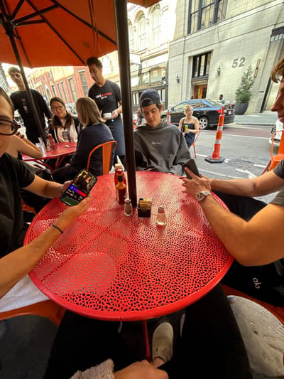 The image depicts an outdoor cafe scene on a sunny day at 52 Temple Place in Boston, Massachusetts, United States.
In the foreground, a bright red circular metal table with an intricate cutout pattern is partially visible. On the table are a ketchup bottle, a hot sauce bottle, two salt shakers, two pepper shakers, and a stack of brown napkins. The hands and forearms of two individuals are visible; one on the left holds a smartphone displaying a video or graphic with a blue wristband on the wrist, while the arm on the right wears a silver watch and holds another smartphone. Below the table, black pants and light-colored shoes are visible.
Several individuals are seated at and around red tables under a large orange patio umbrella. To the left, a young male in a black shirt is partially visible, standing or leaning. A young female with glasses and dark hair is seated, looking right. Next to her, a female with blonde hair in a dark blue quilted jacket is seated with her back partially to the camera. To the right, a young male wearing a backward baseball cap and a grey hoodie is seated, looking down at a smartphone.
A cafe staff member, wearing a black t-shirt with "Café Bonjour Bistro Boston" printed on it and a dark apron, stands between tables, holding a small white saucer.
In the background, a light-colored stone building with "52 TEMPLE PL" inscribed above an entrance is prominently visible, featuring multiple stories and large windows. Parked on the street is a black sedan. An orange traffic cone stands near the curb. Further down the sidewalk, another female in a black strapless top is seated alone at a small table, holding a yellow can.