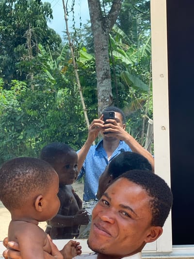 An adult male with dark skin and short dark hair is depicted in a mirror selfie, holding an infant with similar features. The man is smiling at the camera. He wears a light-colored garment on his right shoulder, while the infant is shirtless and looking to the left. The reflection reveals the man holding a black smartphone with multiple camera lenses in both hands to capture the image. In the reflection, he is wearing a blue patterned short-sleeved collared shirt. To the left of the reflected infant, a second shirtless child's reflection is visible. The background, visible in the mirror's reflection, consists of a lush, dense tropical environment under a blue sky, featuring various green trees, large-leafed plants resembling banana trees, and other dense foliage. A white vertical frame element borders the right side of the reflective surface. The scene is located outdoors in Ikot Ete Ekpo, Nigeria.