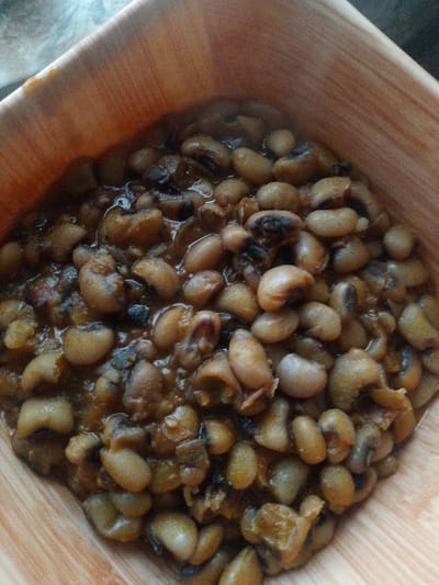 A close-up shot shows a bowl filled with cooked black-eyed peas. The peas are a light tan color with characteristic black "eyes" and are coated in a rich, brown sauce, suggesting they have been stewed or simmered. The texture appears soft and tender. The dish is served in a light-colored, wooden bowl with a visible grain pattern. The bowl's edges are angled, creating a square-like opening. The lighting is somewhat dim, with the light source appearing to be from above and slightly to the left, casting soft shadows. The background is out of focus and appears to be a textured surface, possibly a table or countertop, with a slightly cool tone. The overall impression is that of a home-cooked meal, ready to be served. There is no visible text in the image. The setting implies a casual, domestic environment in Kuru Karama, Nigeria, likely during daylight hours based on the general illumination.