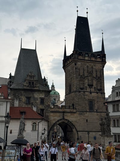 The image depicts the Old Town Bridge Tower in Prague, Czechia, a Gothic tower that stands at the eastern end of the Charles Bridge. The tower is made of stone and features a steeply pitched roof with multiple spires. A large archway, part of the bridge and tower structure, is visible in the center of the image, with people walking through it and along the bridge. Several other buildings with red-tiled roofs and windows are visible to the left and right of the tower. The sky is overcast and cloudy.
In the foreground, a group of people are walking across the bridge. To the left, a street artist is displaying paintings under an umbrella. A statue of a robed figure stands next to a building on the left. To the right of the archway, a multi-figured statue is mounted on a pedestal. The overall scene is that of a busy tourist area, with many people walking and sightseeing.