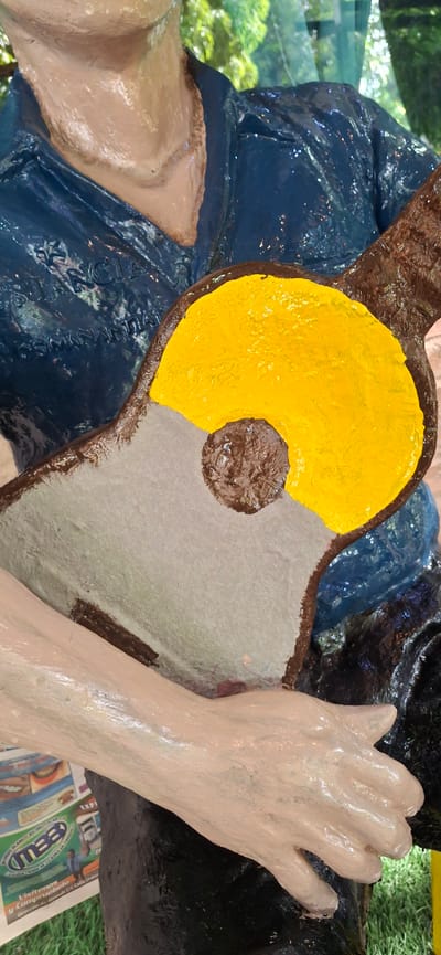 A close-up shot shows a statue of a person playing a guitar in Vallecito, Venezuela. The statue is made of a tan-colored material and wears a shiny blue shirt and pants. The guitar has a yellow soundhole, a brown neck, and a light gray body. The statue's hands are visible, one resting on the guitar and the other holding it. In the background, there is greenery and a newspaper on the ground. The statue is a decorative element in the outdoor environment.