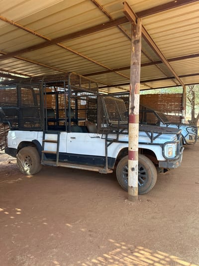 A white, heavily modified pickup truck is parked under an open-sided shelter with a corrugated metal roof. The vehicle features an extensive black metal cage structure that covers the entire truck bed and extends over the cab, indicating it is designed for rugged transport, possibly for safari or utility purposes. A "4x4" emblem is visible on the front of this cage.

The truck is positioned in front of a wooden post, painted with alternating red and white stripes, which serves as a barrier or checkpoint. The ground is reddish-brown, dry dirt or sand, showing tire tracks. Sunlight filters through the roof and sides of the shelter, casting striped and rectangular shadows on the ground and parts of the truck, suggesting it is daytime, likely mid-morning or afternoon.

Beyond the truck, a woven screen or matting is visible, with trees in the background. The shelter's roof is supported by brown metal beams. On the tires, "BRIDGESTONE" and "DUELLER H/T" are visible. A white sticker on the side of the truck's cage contains text that appears to read "PROTECTED AREA," hinting at a location such as a wildlife reserve or restricted zone in rural Senegal. No people or overt activity are depicted; the scene shows a stationary vehicle in a practical, outdoor setting.