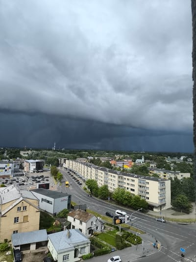 An aerial view captures a dramatic sky over Panevėžys, Lithuania. A dark, heavy storm cloud bank dominates the upper portion of the image, casting a somber mood over the cityscape below. The cloud formation appears textured and layered, suggesting an imminent downpour or ongoing weather event.
The midground features a variety of urban structures. A long, light-colored apartment building with multiple balconies stretches across the frame, its architecture typical of mid-20th century Soviet-era design. Adjacent to it are other buildings, some appearing residential and others more commercial, including a building with large circular signage. A multi-lane road curves through the scene, with several cars and a yellow bus visible, suggesting moderate traffic flow. Trees with lush green foliage line the road and are interspersed throughout the urban landscape, providing a contrast to the concrete and buildings.
In the foreground, smaller residential buildings with pitched roofs are visible. One building has a weathered, rusty metal roof. There are also some smaller outbuildings and gardens. The overall impression is of a densely populated urban area under a menacing sky. The image seems to have been taken from a high vantage point, possibly a tall building or a drone, looking down and across the city. The lighting is dim and diffused due to the storm clouds.