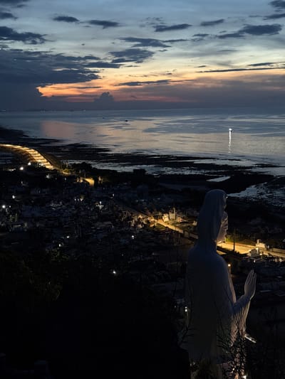 The image captures a twilight scene in the city of Lý Sơn Special Administrative Region, Vietnam. In the foreground, a large, white, abstract statue of a figure is silhouetted against the darkening sky. The statue is facing away from the viewer, with its right arm raised and hand open in what appears to be a gesture of greeting or benediction. The statue is positioned on a dark, overgrown hill, with bare branches of trees partially obscuring its form.
Below the hill and extending towards the sea is a densely populated graveyard. Numerous white gravestones and small structures are visible, illuminated by scattered lights. A road with glowing streetlights winds through the graveyard and along the coastline. The coastline itself is a mix of dark, rocky shorelines and calm water.
The sea stretches out to the horizon, reflecting the soft hues of the sunset. The sky is a gradient of colors, with deep blues and grays at the top, transitioning to oranges and pinks near the horizon, where the last light of the sun is fading. Wispy clouds are scattered across the sky. A few distant boats or buoys are visible on the water, marked by small lights. The overall atmosphere is serene and somber, with a sense of quiet contemplation.