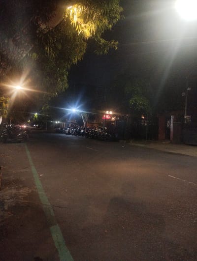 The image captures a night scene on a paved street in Malang, Indonesia. The perspective is slightly elevated, looking down the street, which is dark asphalt with a distinct green line painted along its left edge, indicating a lane or parking demarcation. The street itself appears empty of moving vehicles or pedestrians.
Bright, artificial street lights illuminate the scene from overhead, casting significant lens flares, especially from a powerful light source in the upper right quadrant and another on the far left. These lights create strong glares and a hazy glow in the dark sky. Trees and dense foliage are visible on both sides of the street, with the leaves closest to the light sources appearing bright yellow or green due to the illumination.
Numerous motorcycles and scooters are parked in rows along both sides of the street, suggesting either dedicated parking areas or a busy commercial zone. On the left side, motorcycles are parked beneath the trees and a building facade, while on the right, a denser concentration of parked bikes extends further down the street, obscuring what appear to be low-rise buildings or establishments behind them. Some faint red and yellow lights, possibly from signs or windows, can be seen in the distance behind the parked vehicles on the right.
The overall impression is that of an urban or semi-urban street at night, likely a bustling area given the number of parked vehicles, though it appears quiet at the moment the photograph was taken. No discernible text is visible on signs or buildings due to the low light conditions and distance. The sky is dark, indicating a clear night.