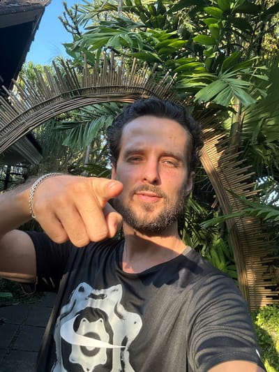 A man with dark hair and a beard points directly at the camera while holding up his right hand. He is wearing a black t-shirt with a white Nike logo and a silver bracelet on his left wrist. The background is filled with lush green palm fronds, a bamboo fence, and a clear blue sky, suggesting a tropical outdoor setting. The lighting indicates it is daytime, possibly late afternoon due to the warm, golden hue on the man's face and the shadows. The overall mood is energetic and engaging, as if the man is interacting with the viewer. The scene appears to be in a tropical outdoor area, with the presence of dense foliage and natural bamboo structures.