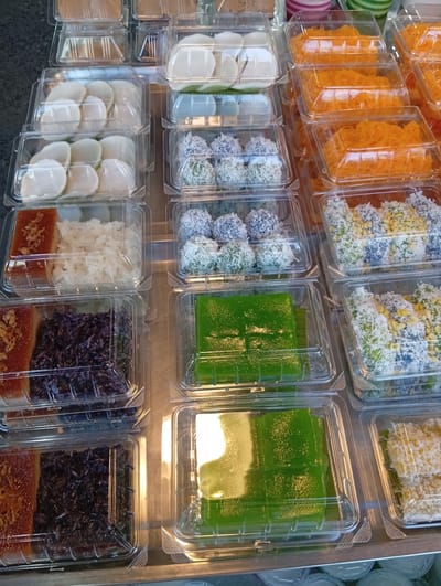 The image shows a close-up, overhead view of a display case filled with various Thai desserts in clear plastic containers. The desserts include sticky rice cakes topped with what appears to be fried shallots, purple sticky rice, green jelly-like cakes, and small, round sweets coated in shredded coconut and other toppings. The desserts are arranged in rows and stacked on top of each other, creating a colorful and tempting display. The setting appears to be an outdoor market or a food stall in Baan Lamai, Thailand, given the context. The lighting suggests it might be daytime. There are no people visible in the frame, but the abundance and variety of desserts imply a busy food retail environment. The containers are neatly arranged, indicating a well-organized presentation for customers. The overall impression is one of a diverse and appealing selection of traditional Thai sweets.