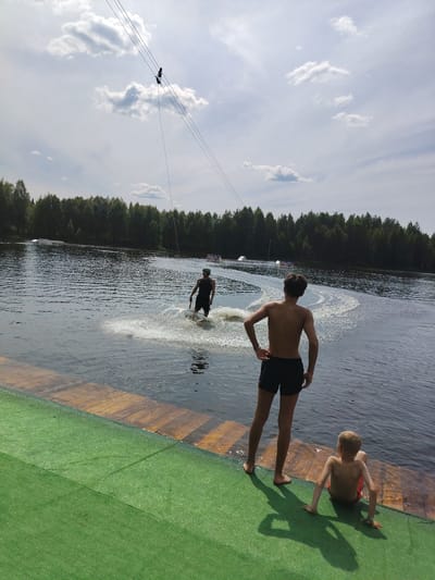 A wide shot shows a wakeboarding facility on a lake. In the foreground, on a green matting, a young boy with blond hair and red swim trunks sits facing the water. To his left, standing and facing away from the camera, is a shirtless teenager wearing black swim trunks. Behind them, on the wooden dock, the water of the lake begins. In the middle ground, a wakeboarder wearing a green helmet and dark shorts is being pulled across the water, creating a spray. In the background, a dense forest lines the far bank of the lake. Above the lake, a cable system is suspended, with a silhouette of a person visible hanging from it. The sky is partly cloudy with scattered white clouds against a light blue background. The scene appears to be in a natural outdoor setting.