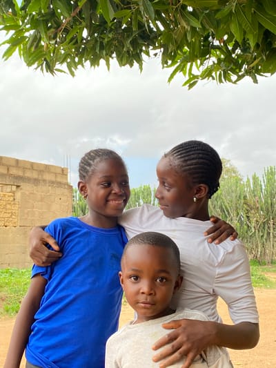 The image shows three individuals in an outdoor setting, likely in Kuru Dungus, Nigeria. The three people, a woman, a girl, and a boy are closely huddled together.
The woman is positioned at the top of the image. She has dark skin, short black braided hair, and is wearing a white shirt. She is looking down towards the other two individuals. The girl, wearing a blue shirt, is smiling and is situated at the bottom of the image. Next to her is a boy in a white shirt, looking at the camera.
The background includes a building constructed from tan-colored blocks and foliage. The sky is partially visible with cloudy conditions. The setting appears to be a rural area. The image seems to be rotated.