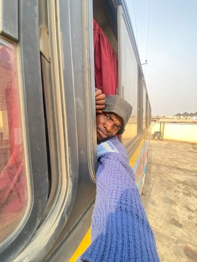 An adult male is observed leaning out of an open window of a stationary passenger bus, positioned on a paved surface in Issele Uku, Nigeria. The individual, of dark complexion, wears a dark bucket hat and a blue knit sweater. He grips the dark-colored window frame with his right hand, his head and shoulders visible from the chest up. His gaze is directed towards the camera with a serious expression. Red curtains are visible inside the bus through the window opening. The bus exterior features yellow and light blue sections. The surrounding environment consists of a bright, clear sky, and in the background, several light-colored multi-story buildings with dark roofs are visible, along with overhead power lines.