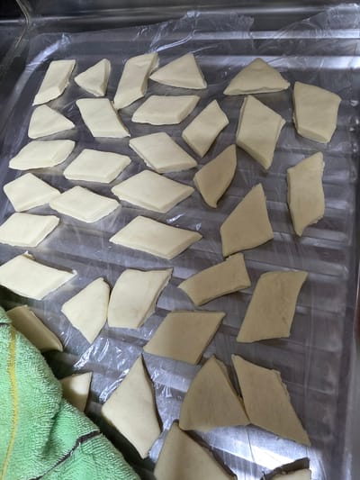 The image shows a collection of raw, light-yellow dough pieces, cut into diamond shapes and laid out on a clear plastic sheet, which is placed on a metallic tray. The dough pieces appear to be of a uniform size and thickness, suggesting they are prepared for a baking or frying process. They are arranged in a somewhat scattered but organized manner across the plastic sheet, filling a significant portion of the tray.
The setting appears to be an indoor kitchen or food preparation area, indicated by the metallic tray and the overall lighting which suggests artificial light or a cloudy day. A green, textured towel with yellow and brown stripes is visible in the lower right corner of the frame, partially covering some of the dough pieces and the plastic sheeting.
No people are visible in the image. The activity taking place is the preparation of dough for cooking, likely for a Korean dish given the location context of Daejeon, South Korea. The dough could be for various items like dumplings, Korean pancakes (jeon), or fried dough snacks. The lighting is neutral, and there are no strong indicators of time of day or weather conditions. There is no visible text in the image.