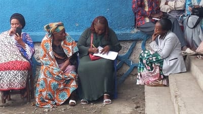 The image depicts an outdoor scene in Goma, Democratic Republic of the Congo, featuring five individuals gathered against a blue-painted concrete wall with faded patterns and a concrete base.

From left to right:
An adult female, wearing a black headwrap and a patterned white, blue, and red dress, is seated on a wooden chair, holding a black smartphone vertically in her right hand. Her gaze is directed towards the right.
Adjacent to her, an adult female in an orange, blue, and brown patterned garment and headwrap is seated, with her body angled slightly towards the right.
The central figure is an adult female with reddish-brown hair, wearing a dark green pleated dress. She is seated on a blue chair and is actively writing on white papers with a yellow pen. A red shoulder bag is visible across her body.
To her right, an adult female in a green floral pleated skirt and a light grey blazer is seated on concrete steps, with her chin resting on her right hand, looking left.
Partially visible behind her on the steps are the lower extremities of a fifth individual.

The ground beneath the subjects is unpaved, appearing as packed dirt with scattered small debris. The overall arrangement suggests an administrative or communal registration process taking place in an informal outdoor setting.