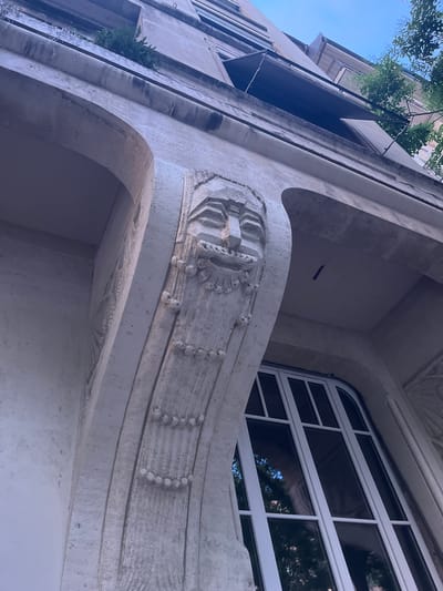 This media file is a low-angle, close-up shot of the exterior of an ornate building, likely an apartment building or historical structure, given the architectural details. The building is made of a light-colored material, possibly stone or concrete, and features curved architectural elements and decorative carvings.

The primary focus is on the building's facade, specifically a balcony or overhang with intricate detailing. A carved relief featuring a stylized floral or geometric pattern is prominent on the balcony's edge. Above this, a large, multi-paned window with white framing is visible, reflecting the surrounding environment, including trees and sky. To the left of the frame, a portion of another building is visible, appearing more modern with vertical window lines. Lush green foliage from trees spills over the edge of the upper structure, suggesting the presence of a rooftop garden or mature trees adjacent to the building.

The scene appears to be set during the daytime, indicated by the bright sky and the reflections in the window. The lighting suggests it could be late morning or early afternoon, with the sun casting subtle shadows. The overall impression is one of historical architectural beauty and integration with nature. There are no people or specific activities depicted, suggesting a still, observational shot. No text is visible in the image. The location context provided is Geneva, Switzerland, which aligns with the elegant and potentially historic architecture.