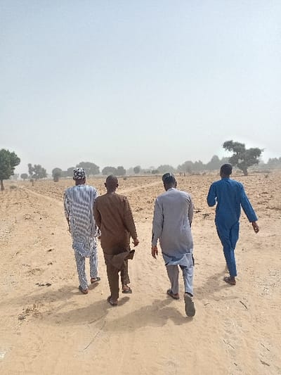 Four adult males are observed walking from a rear perspective along a dusty dirt path in Ayama, Nigeria. The environment is an open, arid landscape under a hazy sky.
The individuals are dressed in loose-fitting traditional attire and open-toed sandals:
- The leftmost man wears a white and blue striped long-sleeved tunic and matching trousers, topped with a patterned cap.
- The second man from the left wears a solid brown long-sleeved tunic and trousers.
- The third man wears a light grey long-sleeved tunic and trousers.
- The rightmost man wears a solid blue long-sleeved tunic and matching trousers.
All four individuals are walking in the same general direction, casting shadows forward and to their left on the light brown earth. The path appears unpaved with visible tracks. The surrounding terrain is characterized by dry, light brown soil with sparse, low-lying vegetation and scattered deciduous trees in the mid-ground and background. The sky is a pale, uniform light grey, indicative of hazy or overcast conditions.