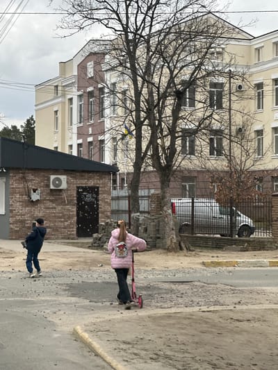The image depicts an outdoor urban scene in Irpin, Ukraine, under an overcast sky. Two individuals, observed from behind, are moving through a street with significant asphalt damage, characterized by potholes and exposed gravel. On the left, a person wearing a dark blue jacket and jeans walks, holding an object. On the right, a person in a pink puffy jacket with a shiny backpack rides a pink kick scooter. In the background, a multi-story building with light yellow/beige and brown sections features numerous windows. A Ukrainian flag (blue and yellow) is affixed to the bare branches of one of the several deciduous trees that partially obscure the buildings. To the left, a single-story brick building with a dark sloped roof has an exterior air conditioning unit and a dark door covered in white markings. A silver van is parked behind a metal fence in the mid-ground. Stacks of grey paving stones are visible near the base of a tree. Utility poles and overhead wires are present against the cloudy sky.