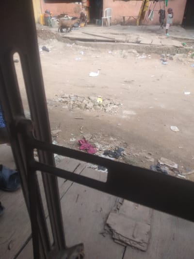 This media file depicts an outdoor scene in Shangisha, Nigeria. In the background, a light-skinned child wearing a striped shirt and pink shorts stands near a clothesline with laundry hanging. Adjacent to the child, a white plastic chair is visible outside a building with an orange-colored wall. Further left, a brown goat stands near some yellow containers and what appears to be a bucket. The ground is dirt, with scattered debris including rocks, plastic bags, and a pink garment. In the foreground, a dark metal frame obstructs the view, suggesting the image was taken from inside a vehicle or through a window with bars. A pair of dark sandals is visible on the wooden floor in the bottom left corner. The overall scene appears to be an everyday setting, possibly a residential area or market, with no specific event or time of day clearly indicated. The lighting suggests it is daytime.