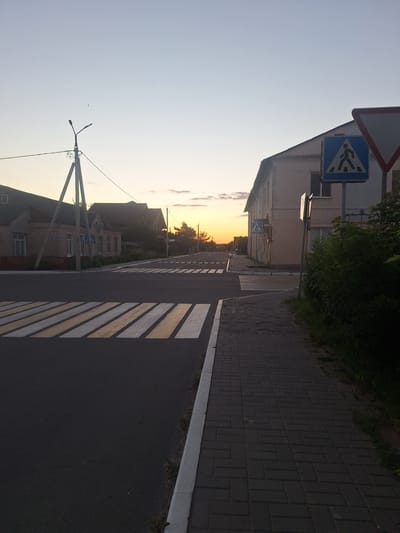 This image depicts a street scene at dusk in the city of Kapyĺ, Belarus. In the foreground, a paved road with prominent white and yellow pedestrian crossing stripes dominates the lower left portion. To the right of the road, a grey brick sidewalk runs parallel, bordered by a white curb. Beyond the sidewalk, dense green foliage is visible.
In the midground, the road intersects with another street. Across the intersection, buildings line the street. On the left side, a brick building with a dark roof and a fence in front is visible, along with a street lamp on a utility pole. On the right side, a lighter-colored, multi-story building stands. A blue pedestrian crossing sign with a white silhouette of a person is mounted on a pole in front of this building. Further down the street, another pedestrian crossing sign is visible on the left.
The background features a clear sky with a gradient from light blue at the top to a pale orange and yellow hue near the horizon, indicating a sunset. Silhouetted trees and smaller buildings are visible against the horizon. The overall impression is a quiet, residential street at the end of the day.