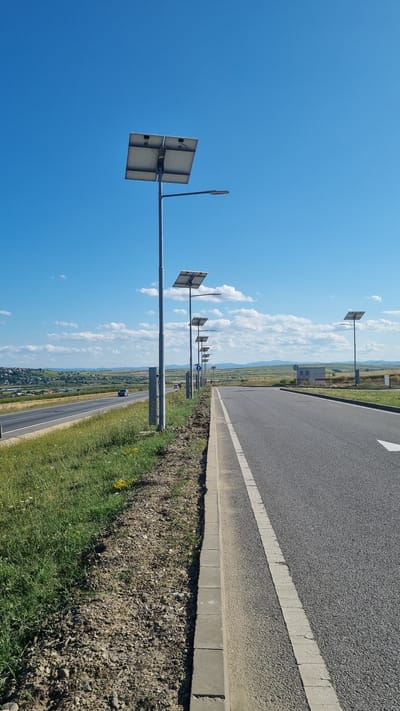 The image depicts a street scene in Șcheia, Romania, characterized by a clear blue sky with scattered clouds. In the foreground, a paved road runs diagonally from the lower right to the upper left of the frame, bordered by a concrete curb and a grassy embankment. A line of solar-powered streetlights with attached solar panels are positioned along the right side of the road, creating a repetitive pattern.

The road itself appears to be a highway or a major thoroughfare, with one lane visible in the foreground and another lane further in the distance. Several vehicles are present on the road, including cars and possibly a truck, traveling in both directions. The surrounding landscape consists of rolling hills covered in greenery, with some buildings visible in the far distance, suggesting a semi-rural or suburban setting. The overall atmosphere is bright and sunny, indicative of daytime.