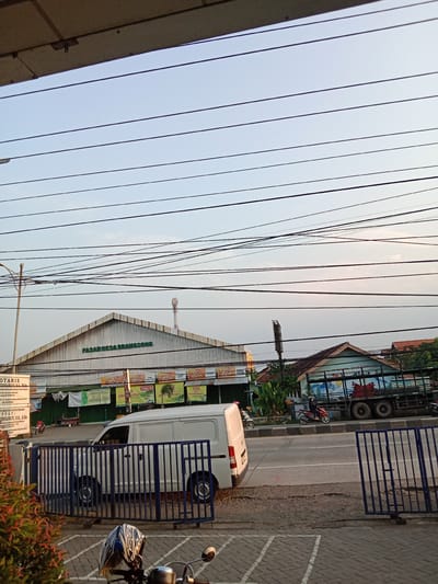 The image shows a street scene in Brangsong, Indonesia, with a large market building in the background. The market, identified by the sign "PASAR DESA BRANGSONG," has a distinctive peaked roof. In front of the market, there are several banners and advertisements, suggesting a vibrant commercial area. A white van is parked on the side of the road, and a blue metal fence runs along the foreground. A motorcycle helmet rests on the handlebars of a visible motorcycle in the lower part of the image. A truck with a loaded bed is also visible further down the road. The sky is clear with a few wispy clouds, and power lines crisscross the scene. The overall impression is of a typical day in a bustling Indonesian town.