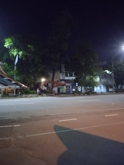 The image shows a street in Surakarta, Indonesia, at night. The street is empty, except for a single person walking in the distance. The road is asphalt and has a slight incline. There are white painted lines running across the width of the road. On either side of the road are trees, bushes, and a building with faded graffiti on its exterior walls. The image was captured from the perspective of a person standing on the sidewalk. The building is located at the coordinates -7.569 latitude, 110.82 longitude. The light is dim and there are street lights in the distance. The image is grainy, which could be a result of low light conditions. There are no cars or other vehicles on the street. The trees obscure the view of the sky, but it appears to be clear. The building on the left side of the road is painted in shades of white and gray, and has a simple, flat roof. There are no visible doors or windows on the building. The building on the right side of the road appears to be made of concrete or brick, and has a more complex facade. The building is decorated with graffiti and faded advertisements, but the details are obscured by the darkness. There are trees on both sides of the street, but they are not in focus. They are all densely packed, suggesting that it is a heavily populated area. The trees are all large and mature. The sidewalk appears to be made of concrete and is wide enough for pedestrians to walk on. There is a long, narrow, gray building behind the trees. The building has a flat roof and seems to be made of concrete. The building has a window on the front and a faded sign that is not visible. The image is taken from a slightly elevated position, which gives the viewer a good view of the street and the buildings. There are no other people visible in the scene. The atmosphere of the image is one of stillness and peacefulness. The lack of activity on the street adds to the sense of calm.