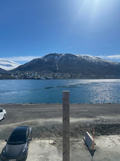 The image shows a scene in Tromsø, Norway, with a view of a body of water (possibly a fjord) and a mountainous landscape. The left side of the image features a mountain range with patches of snow near the top. A town or settlement is located at the base of the mountain along the waterfront.

On the right side, there's a paved area or parking lot. Two cars are visible in the foreground: a dark gray Tesla car and a white car. A wooden beam structure is centrally positioned in the image. A concrete barrier with red and white markings is present on the ground near the water. The sky is blue with a few clouds. The sun is shining brightly, creating a glare on the water surface.