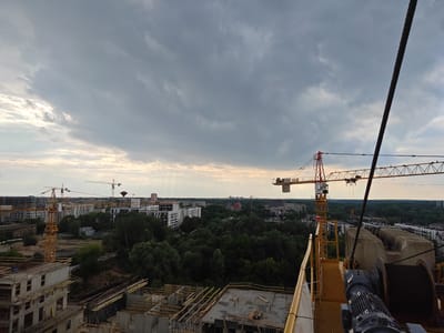 This media file captures an aerial view of a construction site in Poznan, Poland, under a cloudy sky. The scene is dominated by multiple yellow and orange tower cranes in various stages of operation, silhouetted against the overcast sky. In the foreground, unfinished concrete structures of buildings are visible, with wooden scaffolding and rebar indicating ongoing construction. Beyond the immediate construction zone, a dense canopy of green trees separates the development from a cityscape of apartment buildings and residential blocks. In the distance, several taller buildings, including what appears to be a water tower, punctuate the horizon. The sky is filled with heavy grey clouds, with some patches of lighter, possibly sunlit, sky peeking through, suggesting a dynamic weather condition, perhaps the aftermath of rain or the approach of a storm. The overall impression is one of urban development and progress, set against a backdrop of natural greenery. The time of day appears to be late afternoon or early evening, given the soft, diffused light. No people are clearly visible in the frame. A large winch and cable system is prominent in the right foreground, part of the crane's machinery. The image is composed to emphasize the scale of the construction and its integration with the surrounding environment.