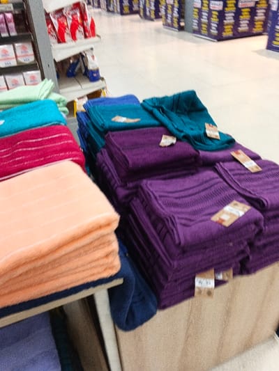 Towels in shades of peach, pink, teal, blue, and purple are stacked on a light-colored shelf in a retail store. Tags are visible on the stacks of folded towels. In the background, other merchandise is displayed on shelves, with the aisle floor a light color. The towels are neatly arranged, showcasing a range of color options.