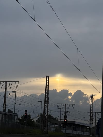 The image captures a view of overhead power lines and train infrastructure against a dramatic sky with a sun pillar effect. The location is a train station in Essen, Germany, likely near the Essen-Altenessen station, given the presence of distinctive overhead line gantries and signaling equipment typical of the area. The sky is overcast, with dark, textured clouds dominating the upper portion of the frame. A bright, vertical column of light, known as a sun pillar, emanates from behind a band of clouds near the horizon, creating a striking visual effect.
In the foreground, a series of high-voltage power lines, suspended from metal poles, crisscross the scene. These poles are part of the railway's electrification system. Below the power lines, a train signal displays a red light, indicating a stop. A covered platform area is visible to the left, with a dark glass facade. Trees and other vegetation are sparsely present along the ground.
The overall atmosphere is somber, with the dramatic sky and the utilitarian nature of the train station creating a sense of industrial realism. The presence of the sun pillar adds an element of natural phenomenon to the otherwise man-made environment. No people are clearly discernible in the image. The composition is dominated by the interplay of man-made structures and atmospheric conditions.