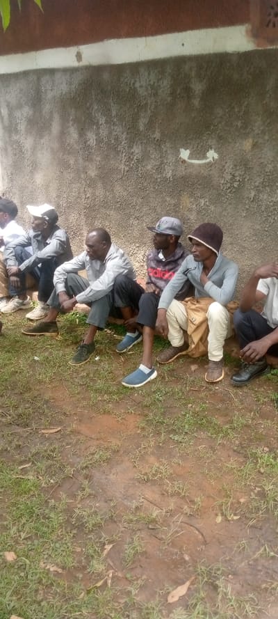 The image shows a group of five men sitting in a line on a dirt path in front of a concrete wall in Kivaywa, Kenya (latitude: 0.56, longitude: 34.806). The men are all wearing casual clothing and footwear. They are all looking in different directions. There is a small amount of green grass growing on the path. The concrete wall is cracked and weathered.  There appears to be a white arrow painted on the wall.