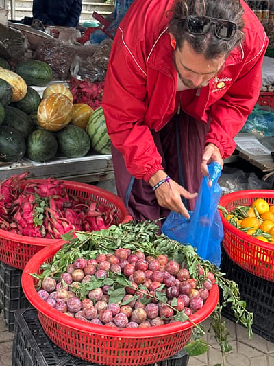 A man wearing a red jacket over a dark maroon garment is at a market stall in Bắc Yên, Vietnam, with an array of fruits and produce. In the foreground, a large red plastic basket is filled with ripe red plums and some green leaves. To the left of the plums, another red basket overflows with red dragon fruit. Further back, in a white plastic tray, are various melons, some green and striped, others yellowish with lighter stripes. Behind the melons, there are bags of what appear to be dried goods. The man is leaning forward, holding a blue plastic bag open with his left hand and reaching into the basket of plums with his right, indicating he is selecting or bagging the fruit. He has sunglasses perched on his head and wears a bracelet on his left wrist. The ground is paved with stone tiles.