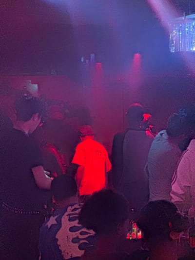 The image depicts an indoor nighttime gathering, likely at an event such as a concert or club, illuminated by striking artificial red and purple-blue lighting. Several individuals are present, situated in a dynamic and dimly lit environment.
In the foreground, a person is visible from the waist down, holding a small device, possibly a phone, with both hands. Adjacent to them, another individual wears a dark garment featuring a distinctive white and blue flame-like pattern with a star design on the lower back or leg area. Nearby, a person is seen from the back, wearing a bright red t-shirt. On the back of this red shirt, near the collar, there is some white or light-colored stylized text, possibly a logo, arranged in a curved or circular fashion; however, the specific words are illegible due to the image resolution and lighting conditions.
Further into the background, additional figures are visible, some standing or leaning, their features largely obscured by the low light and shadows. One person appears to be wearing a light-colored shirt or jacket over a darker one. Streaks of bright, possibly digital, light sources are visible in the upper left corner of the frame, contributing to the vibrant and energetic atmosphere of the event. The scene suggests an active social gathering taking place in Linghe, China, characterized by its intense artificial illumination.