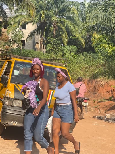 Two women rest on a yellow vehicle, possibly a taxi or van, in a tropical setting. One woman, wearing a light blue top and jean shorts, props herself up while the other, in a black tank top and jeans, lies seemingly asleep with a purple floral top beside her head. Behind them, a man sits on a motorbike, partially obscured. Lush green palm trees and foliage surround the scene, with a glimpse of a building in the background. The ground appears reddish brown.