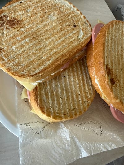 The image shows a close-up view of three toasted sandwiches on a white plate and a textured white napkin. The sandwiches are made with sliced bread that has been grilled, as evidenced by the prominent grill marks. The sandwiches appear to be filled with cheese and some kind of pink processed meat, possibly bologna or mortadella. A thin layer of white spread, likely mayonnaise or cream cheese, is visible on one of the sandwiches. The napkin beneath the sandwiches has a black outline drawing of what appears to be a cloud or a map of a country. The background is a neutral surface, likely a countertop or table. The lighting suggests an indoor setting. There are no people or any indication of the specific location within Vratsa, Bulgaria.