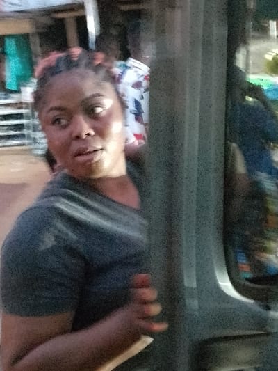 The image depicts a partial view of a female subject, likely inside a vehicle, observing an external environment in Orie Uratta, Nigeria. The subject, a dark-skinned female with reddish-highlighted braided hair, is positioned on the left side of the frame, visible from the chest upwards. She wears a dark gray t-shirt and exhibits an attentive expression, with her gaze directed towards her left (viewer's left) and her mouth slightly agape. Her right hand is seen gripping a prominent vertical gray pillar or frame, consistent with a vehicle's interior structure, which vertically bisects the right half of the image. The background is out of focus, revealing an outdoor setting consistent with a street market. Indistinct figures are discernible, including one partially visible figure wearing a white patterned garment. Blurred objects resembling merchandise or market stalls, such as a turquoise item and metallic shelving, are also present. The scene is illuminated by subdued lighting.