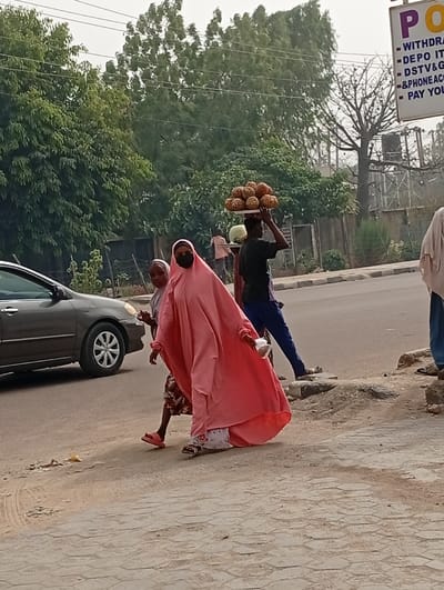 This image captures a street scene in Darmanawa, Nigeria. In the foreground, a woman wearing a long, flowing pink abaya, black face mask, and sunglasses walks towards the viewer's left. She holds a small white object in her right hand. Adjacent to her and slightly behind, a shorter individual, possibly a child, in a patterned top and pink sandals, is also walking. In the midground, a young male individual wearing a dark t-shirt and blue pants walks right to left, expertly balancing a flat white tray loaded with numerous spherical, spiky fruits, likely pineapples, on his head. Further back and to the right, another individual is partially visible, also appearing to carry an item on their head. On the far right, a partial view of an individual's lower body in light-colored clothing and blue pants is seen.
The environment includes a paved road on the left, where a dark-colored sedan is partially visible. A dirt shoulder separates the road from a patterned hexagonal-tiled sidewalk where the individuals are walking. In the background, lush green trees and some bare tree branches are visible under an overcast sky. Overhead power lines traverse the upper portion of the scene. On the far right, a commercial sign with purple lettering is partially visible, displaying text such as "WITHDRA," "DEPO IT," "DSTV&G," "&PHONEACC," and "PAY YOU," suggesting a point-of-sale or payment service business. The scene depicts typical street activity with vendors and pedestrians.