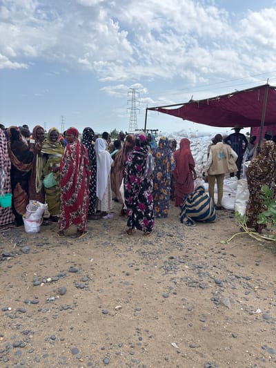 The image depicts a group of people, predominantly women, gathered outdoors in what appears to be a distribution or aid site. The ground is covered with gravel and sand. In the background, several large white sacks are stacked under a burgundy-colored tarp, suggesting they contain supplies. Tall electricity pylons are visible in the distance, and the sky is partly cloudy. Some individuals are wearing colorful traditional clothing, while others are in more neutral attire. One person in the foreground, wearing a vest with a logo, appears to be an aid worker. The scene suggests a humanitarian aid distribution or a market setting where goods are being exchanged. The specific location in Ad-Damazin, Sudan, is not explicitly identifiable from the visual cues alone, but the presence of a large group of people in an outdoor setting with stacked sacks of goods is consistent with aid operations in regions facing hardship.