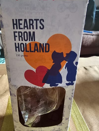 A sideways close-up shows a box of "Hearts from Holland" chocolates, weighing 150 grams. The box is white, and features an orange, red, and blue graphic of a couple. The words "Hearts from Holland" are printed in large, bold, dark-blue letters. An opening in the box displays a clear plastic bag filled with the chocolates. The box is placed on a light-brown or beige surface, possibly a couch or similar furnishing. The background is out of focus, showing darker fabric colors. No people or specific location details (such as Holywell, United Kingdom) are visible in this image.