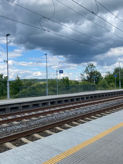 The image shows a view of a train station platform in Lahošť, Czechia, under a cloudy sky. In the foreground, there are train tracks with ballast and concrete ties. The platform itself is paved with hexagonal tiles and features a tactile warning strip along the edge. Beyond the tracks, a metal fence separates the platform from a grassy area and trees. Several lampposts and overhead power lines for the trains are visible. In the background, rolling hills covered in green vegetation and trees are seen, along with a few distant structures. A sign indicating "1A" is mounted on a pole on the platform. There are no people visible in the image.