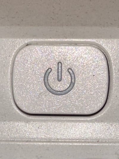 A close-up shot shows a power button on a light-colored electronic device. The button is round and slightly indented, with the universal power symbol imprinted on it. The symbol is a circle with a vertical line extending from the top, indicating the power on/off function. The surrounding surface of the device has a textured, speckled appearance. There is a small dark speck on the surface near the symbol. The lighting is even, highlighting the texture of the material. The image does not provide any information about the location in Melitopol, Ukraine.