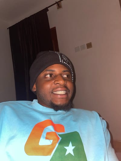 The scene depicts a male individual positioned indoors, located in Ube, Nigeria. He has a dark complexion, a short beard, and is wearing a black knit beanie embellished with silver reflective studs on the right side. His attire includes a light blue long-sleeved sweatshirt featuring a prominent graphic of an orange 'G' partially overlaid by a green shape containing a white star. The individual's head is turned slightly to the right, with his eyes directed towards the right side of the frame and upward. He exhibits a wide smile, revealing white teeth. The background features a plain light-colored wall. Dark brown curtains hang from a rod on the left side of the frame. On the wall to the right, three rectangular elements, consistent with electrical outlets or switches, are visible in white, light brown, and darker brown finishes.
