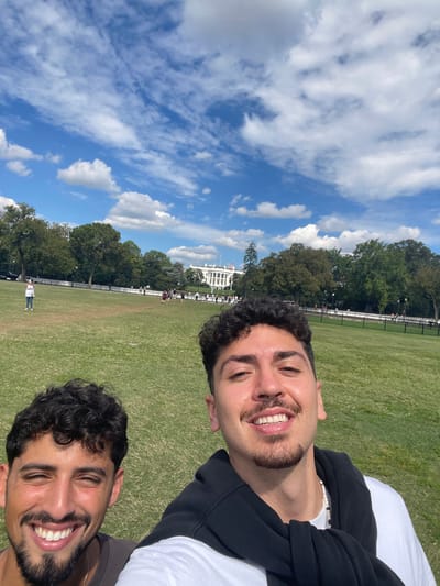 Two young adult males are featured in a selfie-style photograph. The male on the right, centrally positioned and smiling broadly with visible teeth, has dark curly hair, a mustache, and a chin-strap beard. He wears a white t-shirt with a black sweatshirt draped over his shoulders and a white beaded necklace. The male on the left is partially framed, smiling with visible teeth, possessing dark curly hair and a beard, and wearing a dark t-shirt.
They are situated on a large, vibrant green grass lawn. In the midground, a dark security fence delineates the boundary of the lawn, behind which numerous indistinct individuals are visible. Further in the background, a dense line of green trees partially frames the iconic white neoclassical building of the White House, distinguished by its columns and a flag flying on the roof. The sky above is bright blue with scattered white cumulus clouds.
The image was captured in Washington, D.C., United States, with the subjects positioned on a public park area, likely the Ellipse, facing the South Lawn and South Portico of the White House. The scene depicts a clear, sunny day.