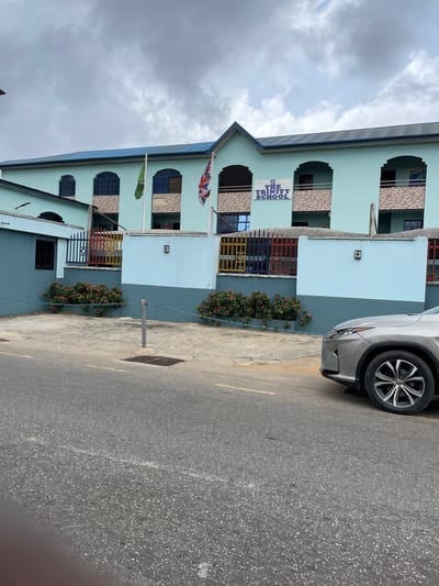 The image shows a multi-story building identified as "The Trinity School," likely located in Ikeja, Nigeria, based on architectural style and the presence of a car bearing a Nigerian license plate. The building has a light blue exterior with dark-framed windows and balconies decorated with colorful railings. A British flag and another green flag are visible. A silver car is parked in front of the building, partially obstructing the view. The street in front of the building is paved, with a manhole cover visible. The sky is overcast.