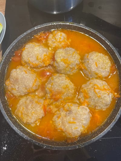 A close-up, overhead shot shows eight meatballs in a shallow, orange-colored sauce within a dark-grey, speckled frying pan. The meatballs are roughly spherical and appear to be made of ground meat and possibly rice or breadcrumbs, with a slightly textured surface. Thin strands of grated orange carrot are visible sprinkled over and around the meatballs, along with some diced red tomato in the sauce. The frying pan sits on a black, flat stovetop. To the left of the pan, a sliver of a light blue plate with a yellow circular object is visible. The overall scene is well-lit, with the focus on the food. There are no people, animals, or elements of the urban environment of Chelyabinsk, Russia, present in the image.