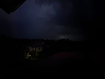 The image captures a nocturnal outdoor scene in Zornitsa, Bulgaria, under a heavily overcast sky. The sky displays a dark blue-purple hue, with a subtle, diffuse bright area visible in the upper right quadrant, consistent with a distant lightning flash illuminating the cloud cover.
In the upper left foreground, a dark, angled architectural element, likely an overhang or roof eave, partially intrudes into the frame. The middle ground on the left features a single-story house with a light-colored facade and a dark, sloped roof, illuminated by a solitary, bright artificial light source positioned at ground level nearby, casting a warm glow on the immediate surroundings. Further into the distance, scattered faint artificial lights delineate the presence of other structures or a distant settlement against the dark horizon. The lower right foreground reveals a partial view of a reddish-brown roof belonging to another structure. No individuals, animals, or discernible activities are present within the scene. The overall environment suggests a dark, potentially stormy evening in a residential or suburban area.