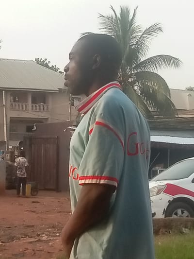 An adult male stands in profile, facing left, in an outdoor setting in Ogidi, Nigeria. He possesses dark skin, short dark hair, and a visible beard or stubble, with his mouth slightly ajar. He wears a light blue short-sleeved polo shirt featuring red and white striped accents on the collar and sleeve cuffs. Red lettering, partially visible as "GIG," is present on his right sleeve.
Behind the subject, to the left, a multi-story building with a grey corrugated metal roof and balconies is visible. Further left, a second individual, a smaller figure, is observed standing near a brown wooden gate, adjacent to a yellow container and a wheelbarrow on reddish-brown earth. To the right of the main subject, a large palm tree with prominent fronds extends upwards. A white vehicle, possibly a compact SUV, with a distinctive red stripe along its side, is partially visible on the far right, parked on or beside the unpaved ground. The immediate foreground consists of reddish-brown dirt, with a small patch of green grass on the extreme lower right. The sky is a uniform, light grey, indicating overcast conditions or haze.