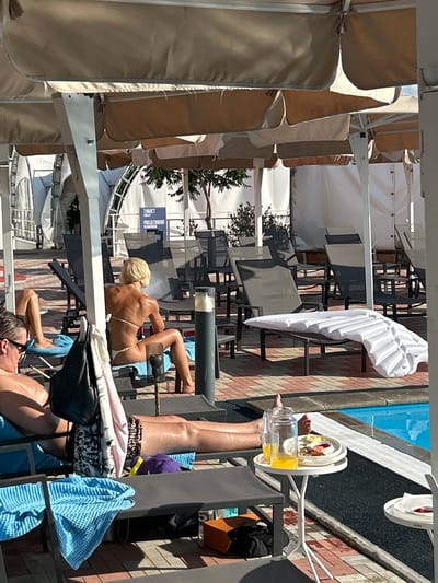 This image depicts an outdoor scene at what appears to be a resort or pool area in Novokuybyshevsk, Russia. In the foreground, several lounge chairs are arranged on a brick-patterned walkway adjacent to a blue swimming pool. Some chairs are occupied by individuals sunbathing. One person, a woman with blonde hair, is seated on a lounge chair, facing away from the camera, wearing a white bikini. Another person, seen from the waist down, is lying on a lounge chair with a purple hat and patterned shorts. A small white table stands beside one of the chairs, holding drinks and a plate of food. In the background, more lounge chairs and tables are visible under a series of large, beige umbrellas. Signs with text in Cyrillic are also present, one appearing to indicate restrooms. The overall environment suggests a sunny day at a recreational facility.