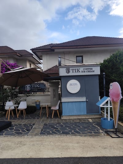 The image shows an outdoor seating area for a cafe and ice cream shop in Lâm Viên - Đà Lạt, Vietnam. A large beige umbrella shades a circular table with two white chairs. To the right, two more white chairs are arranged around a small table. A sign above the outdoor seating reads "TIK RESTAURANT COFFEE". A grey kiosk-style building is visible behind the seating area, with a prominent sign that says "TIK COFFEE ICE CREAM". A circular sign on the kiosk reads "BUDS ICE CREAM OF SAN FRANCISCO". To the right of the kiosk is a large cutout of a pink ice cream cone. The ground is paved with dark hexagonal and rectangular stones. In the background, there are multi-story houses with tiled roofs and lush green trees. The sky is partly cloudy.
