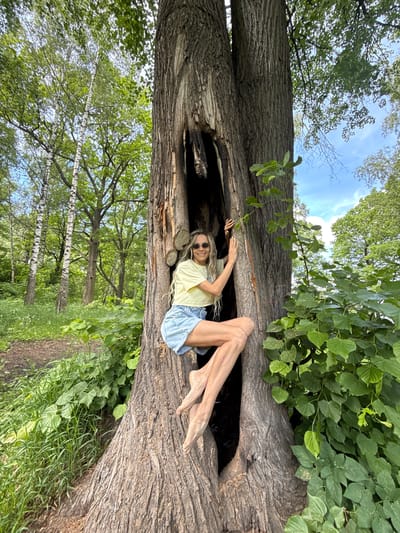 A young woman with long blonde dreadlocks, wearing sunglasses, a light yellow t-shirt, and denim shorts, is lying inside a large, hollowed-out fallen tree trunk in a forest. She is smiling broadly and looking towards the camera, with her arms raised and hands resting on the inside of the tree. Her bare legs extend out of the hollow. The tree trunk, characterized by rough bark, dominates the foreground, running diagonally across the frame.
The surrounding environment is a lush green wooded area, indicative of daytime with bright sunlight. Above the tree, a clear blue sky with some white clouds is visible through a canopy of green leaves from various trees. To the left and in the background, numerous trees with dense green foliage are present, including distinct birch trees with their characteristic white bark. A dirt path or trail can be seen winding through the grass and undergrowth in the lower right background. The scene suggests a relaxed and joyful atmosphere. There is no visible text in the image. The location context is Rusanovo, Russia.