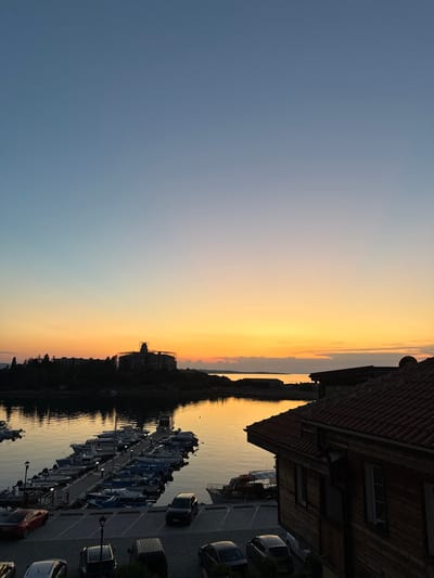 The image depicts a waterfront scene at sunset in Sozopol, Bulgaria. The sky transitions from a deep blue on the left to a vibrant orange and yellow near the horizon, indicating the time of day is either sunrise or sunset. The calm water reflects the warm colors of the sky and the silhouettes of buildings and trees along the opposite shore.
In the foreground, a row of moored boats of various sizes lines a dock. Several cars are parked along a paved area next to the boats. A two-story building with a tiled roof and wooden siding is visible on the right, with windows and architectural details suggesting it might be a residence or commercial establishment.
The overall mood of the image is peaceful and serene, characteristic of a quiet evening by the sea. The warm lighting creates a tranquil atmosphere. There is no visible text in the image.